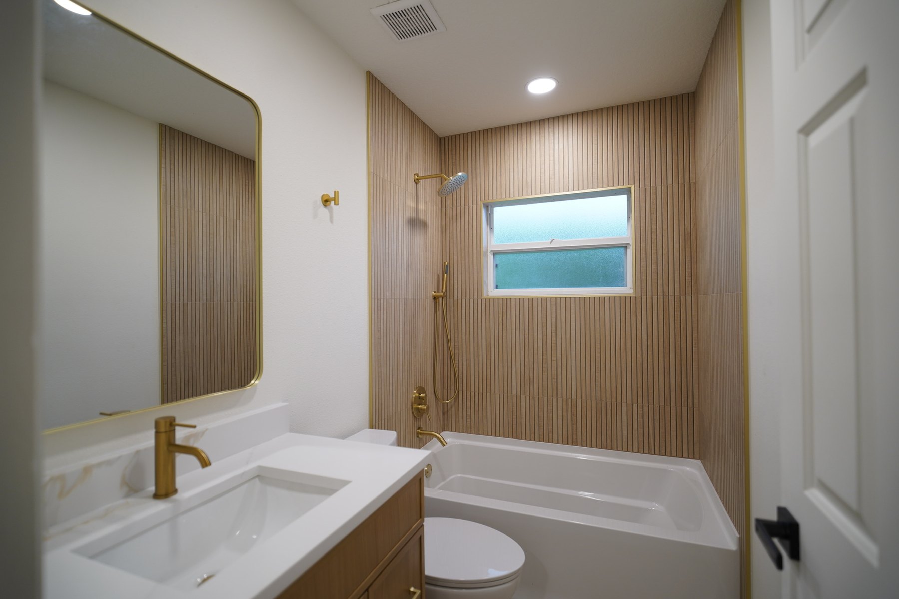 The same bathroom finished: a new tub in a vertical wood-look tile surround, brass shower fittings, a new vanity with a stone top and a brass-framed mirror.