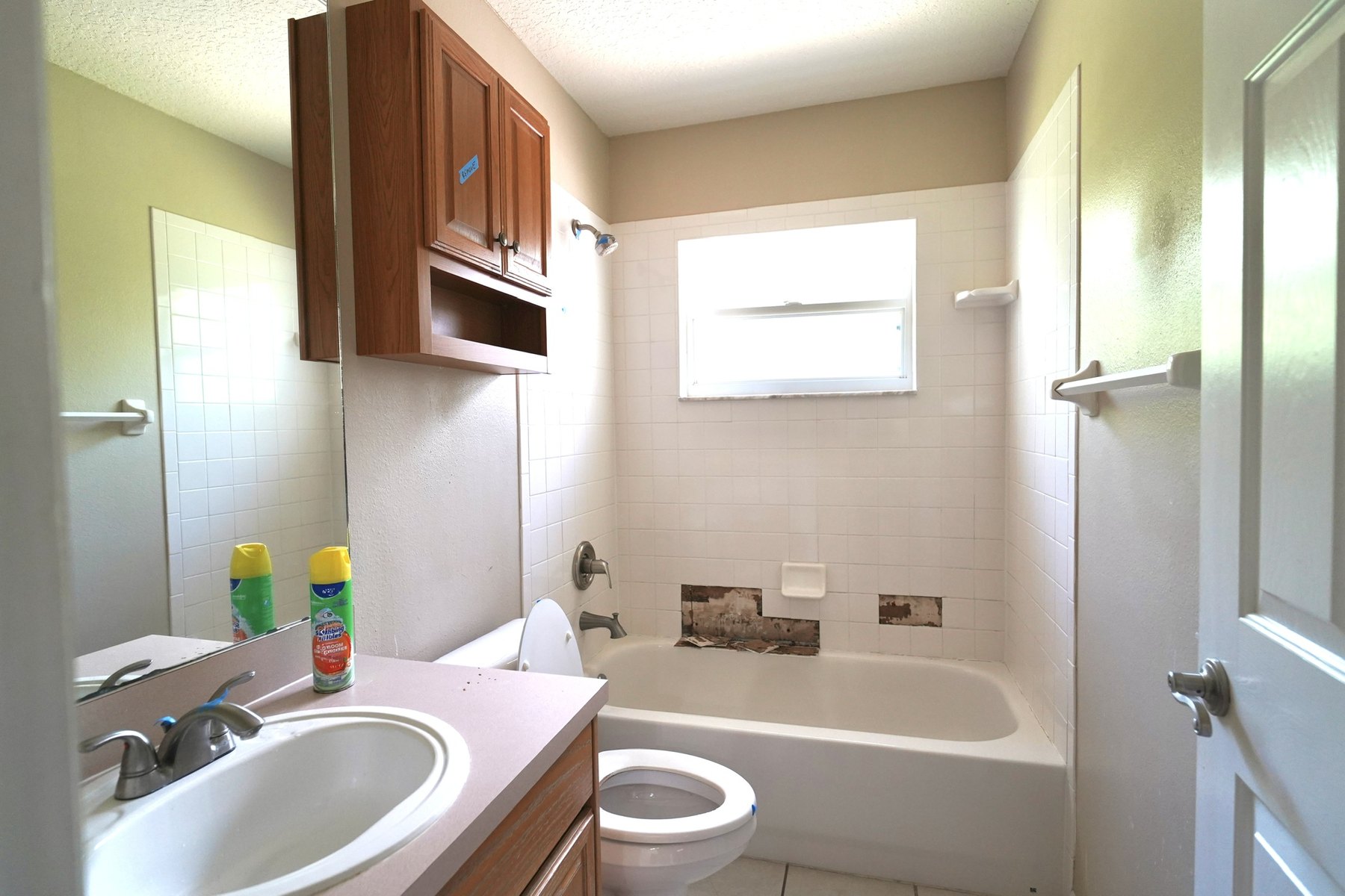 A tub bathroom before the remodel: white tile surround with a small window, a laminate vanity with an oak overhead cabinet, green walls.