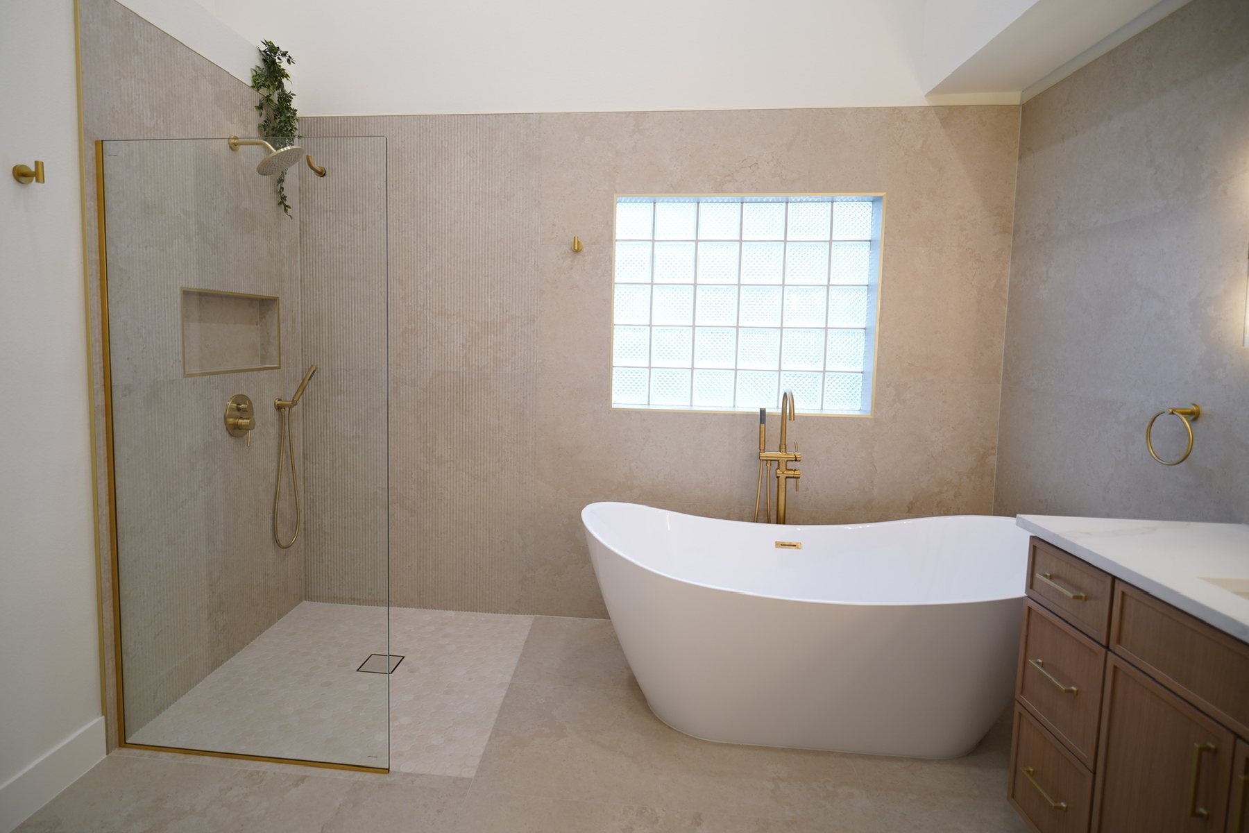 The same primary bathroom finished: an open tiled shower behind a glass panel, a freestanding tub under the same glass-block window, and a wood double vanity.