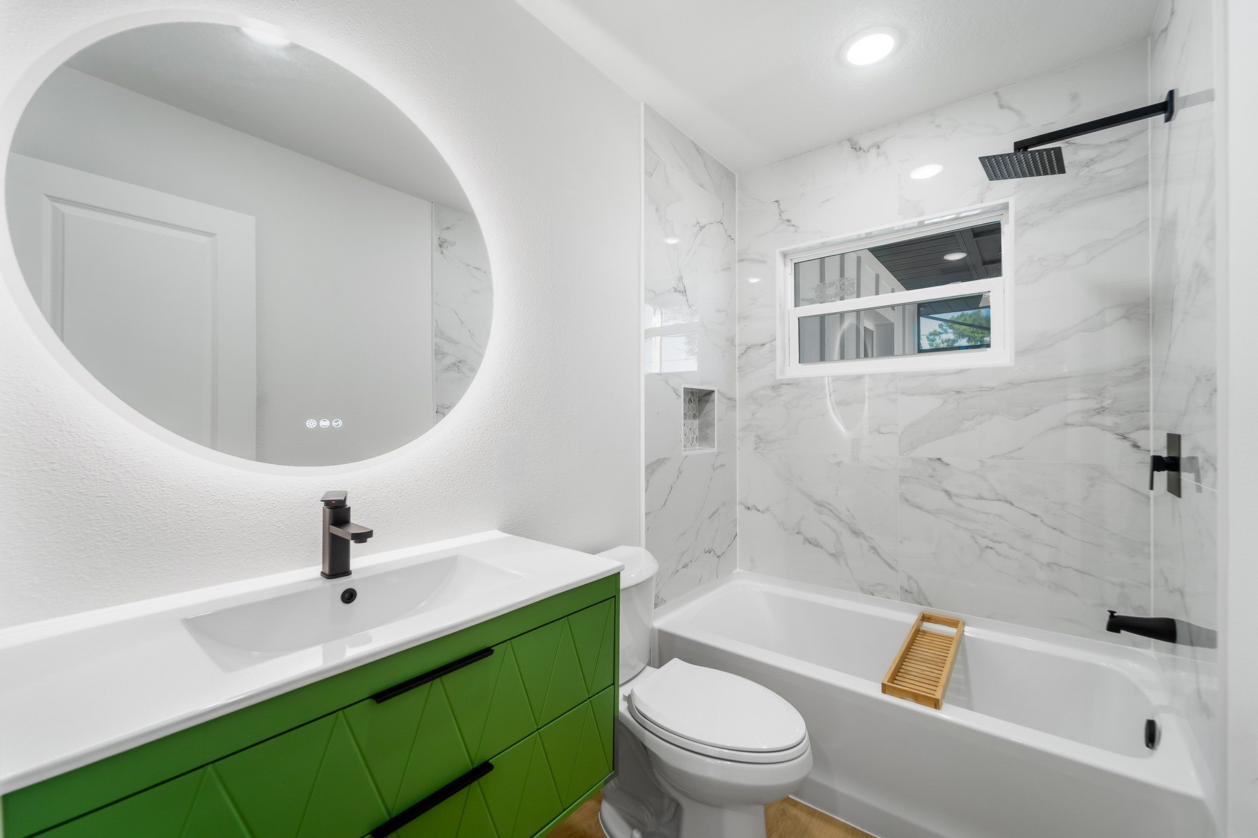 The same bathroom finished: a new tub in marble-look tile, a rain head and matte black fittings, a green vanity and a round lit mirror.