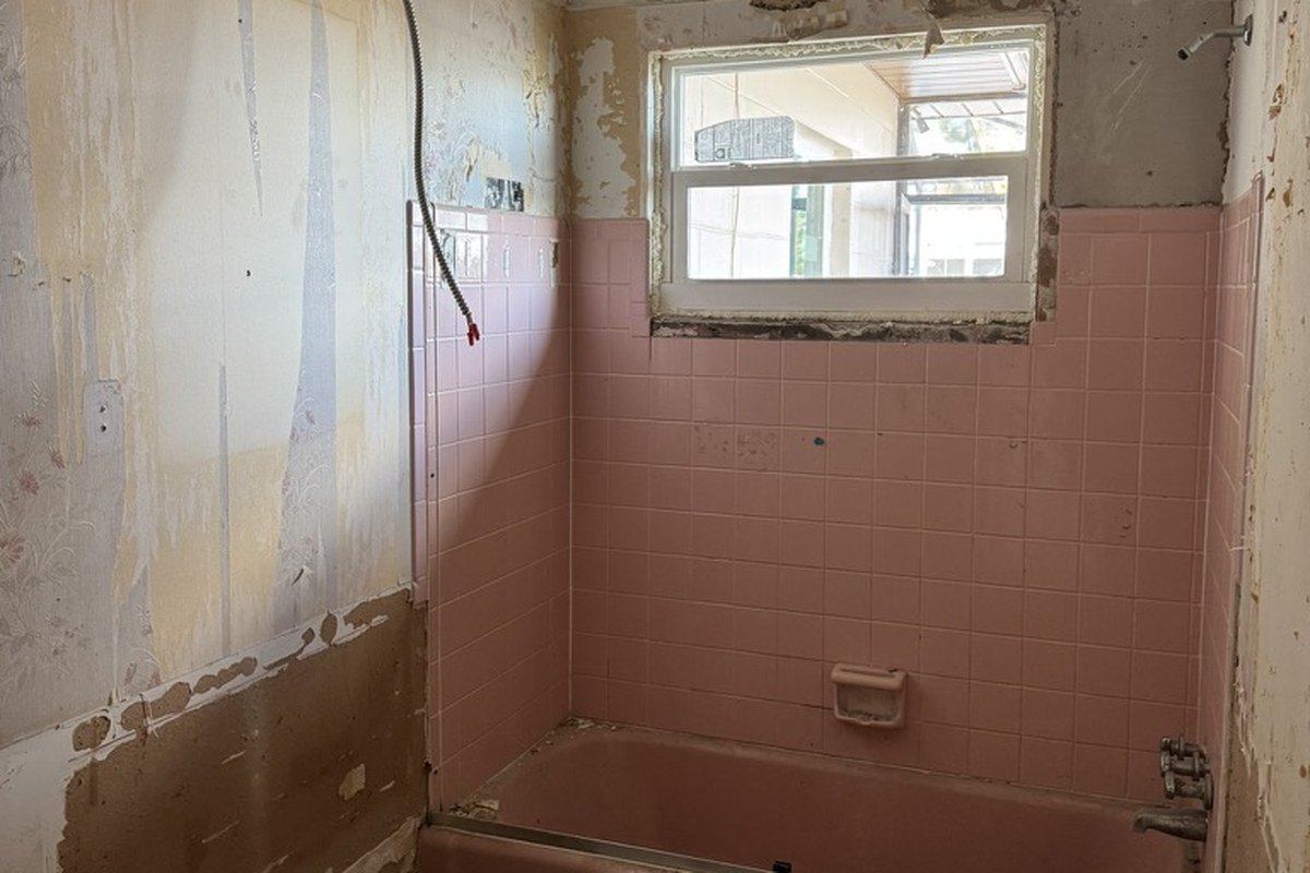 The bathroom stripped for the remodel: pink tile around a bare tub, walls scraped to the plaster, the old window still in the tub wall.