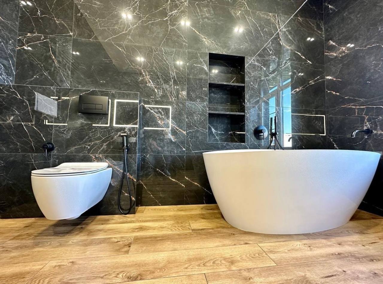 The same dark marble bathroom from the tub side: a white freestanding tub, a wall-hung toilet, black fittings and a lit shelf niche.