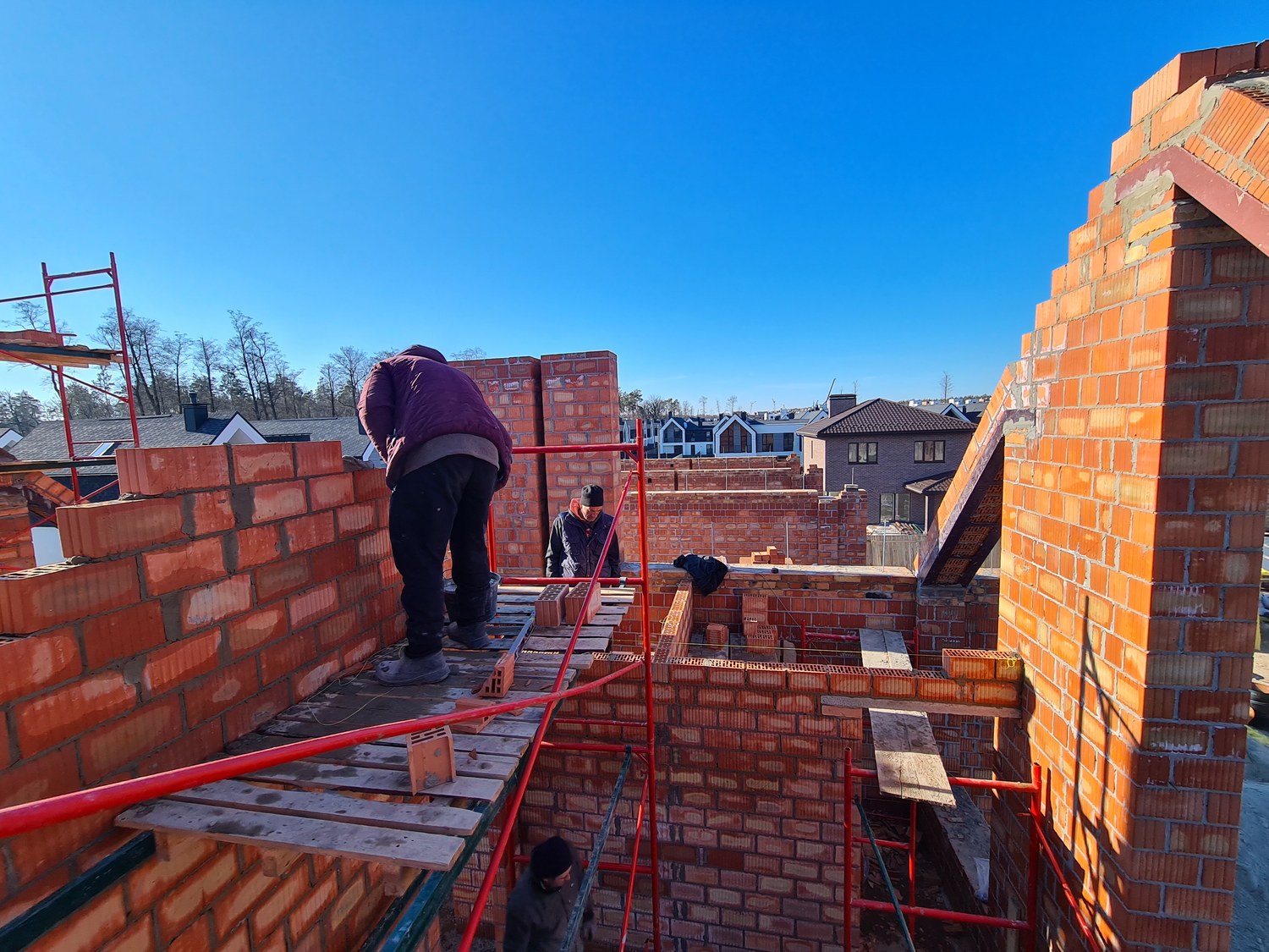 Masons laying brick gable walls at height on scaffolding, mid-construction townhouses in the background.