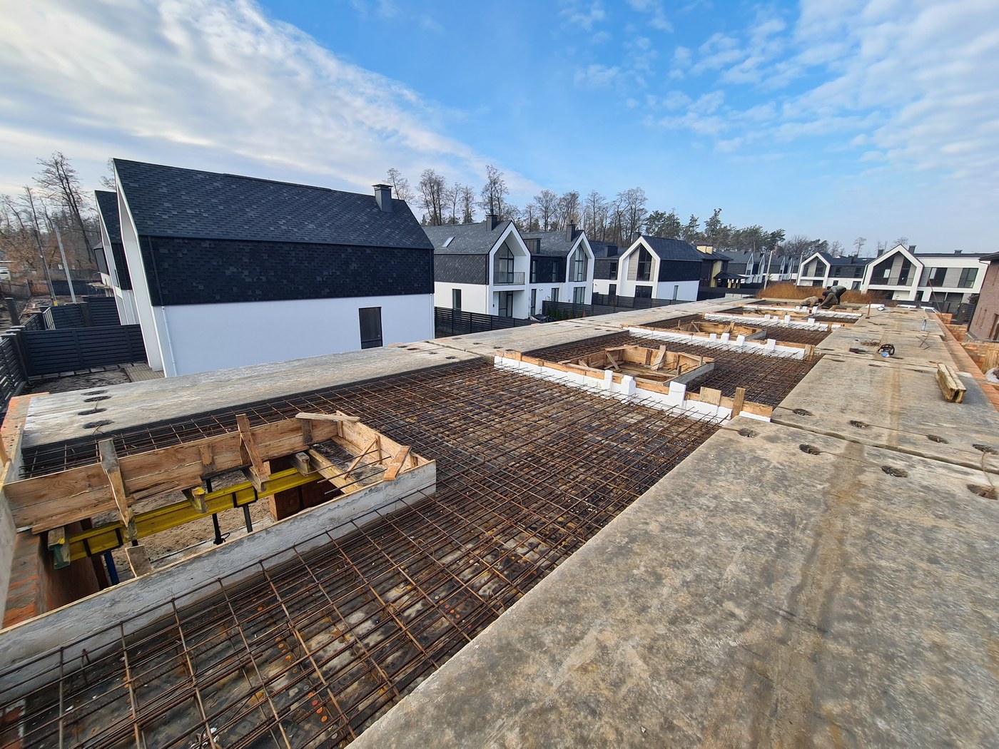 Elevated view of rebar and formwork for a second-floor slab, with a row of completed townhouses standing in the background.