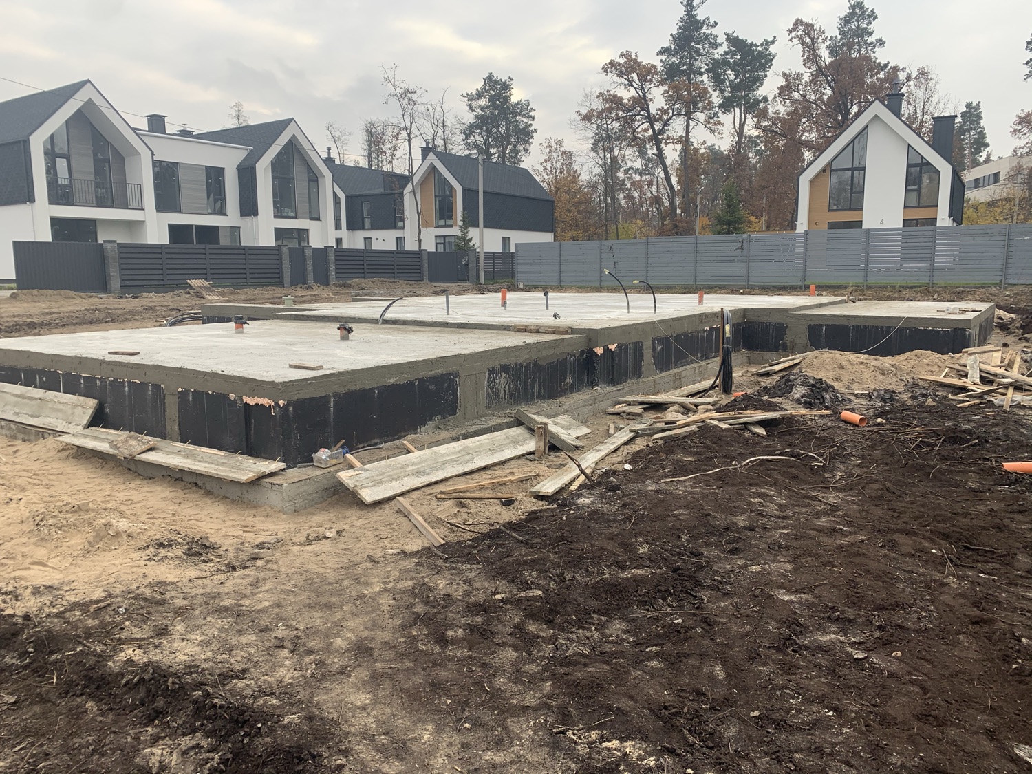 Completed foundation with waterproofing membrane and rebar, ready for masonry, neighboring finished houses visible behind.