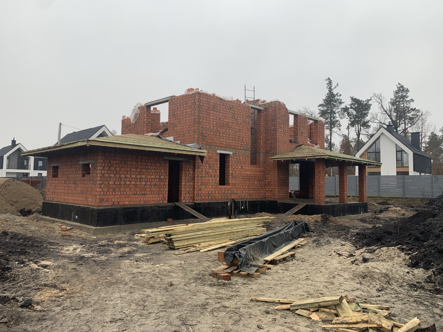 Brick masonry shell with hip-roof timber framing complete, covered porch structure framed.