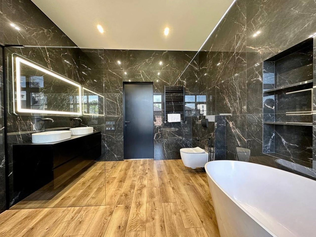 Primary bathroom finished in book-matched dark stone, with a double vanity, freestanding tub, and backlit mirror.