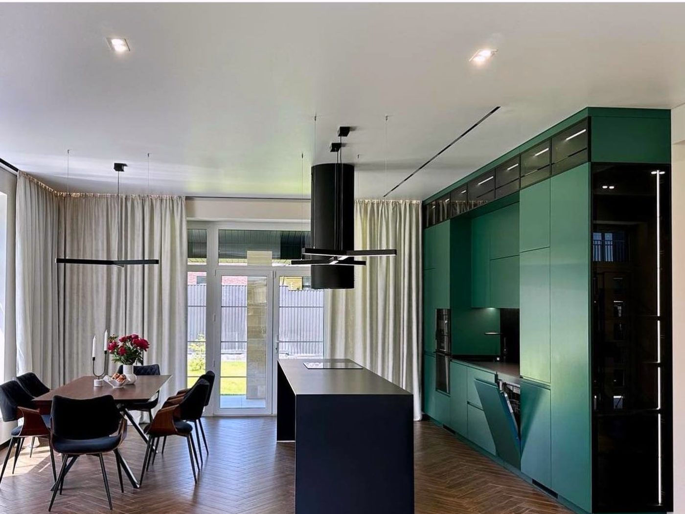 Open living space with emerald full-height cabinetry, a black island, herringbone floor and a dining table by the window.