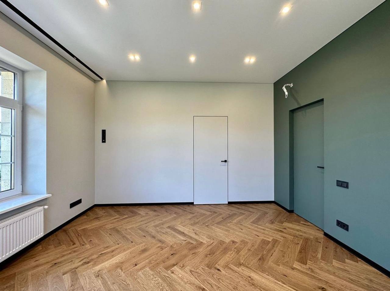 A finished room with a herringbone wood floor, white walls, recessed lights and a green feature wall with a flush concealed door.