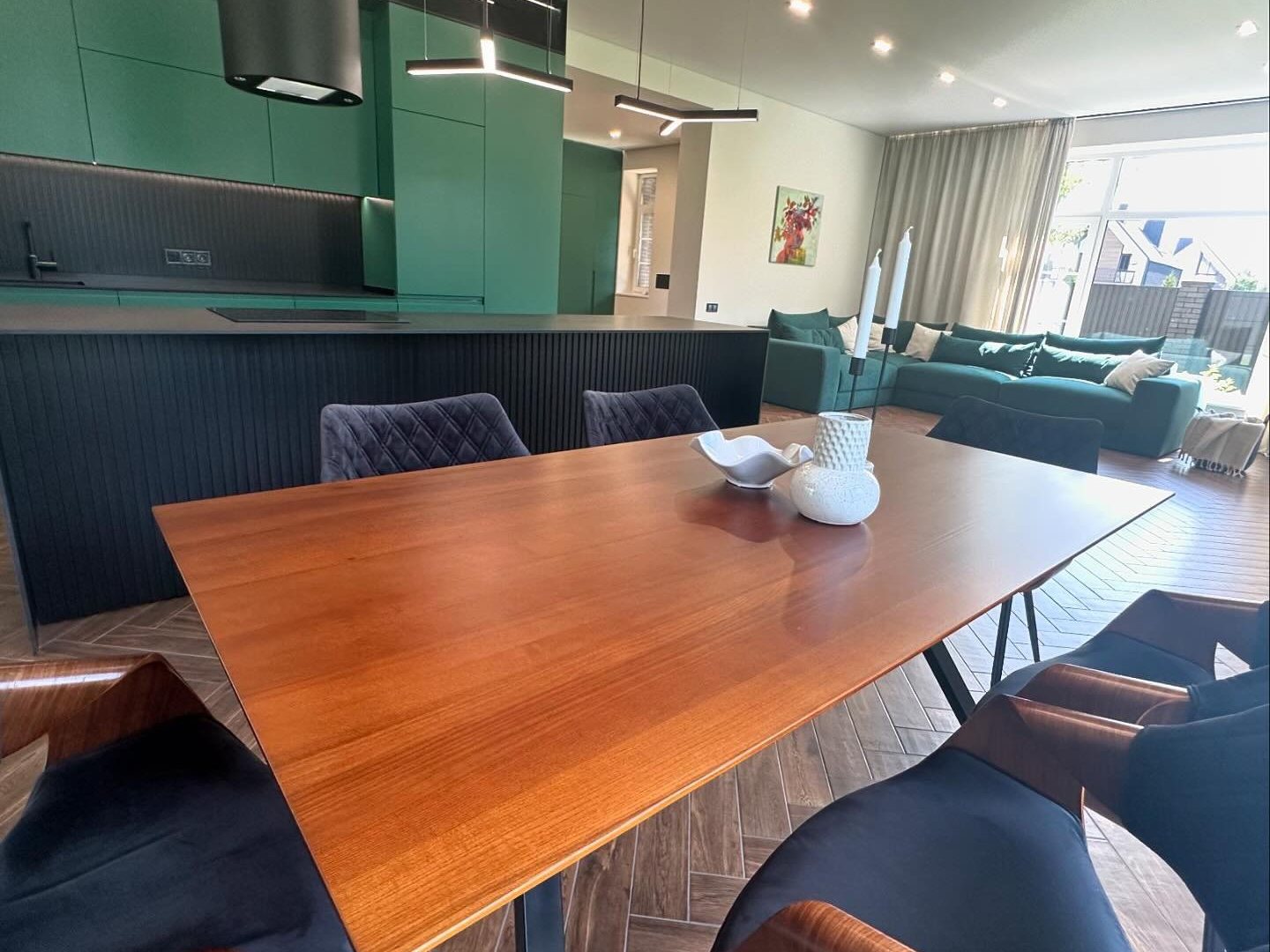 The dining table in the same open room, the black kitchen island and green cabinetry behind it and the living area beyond.