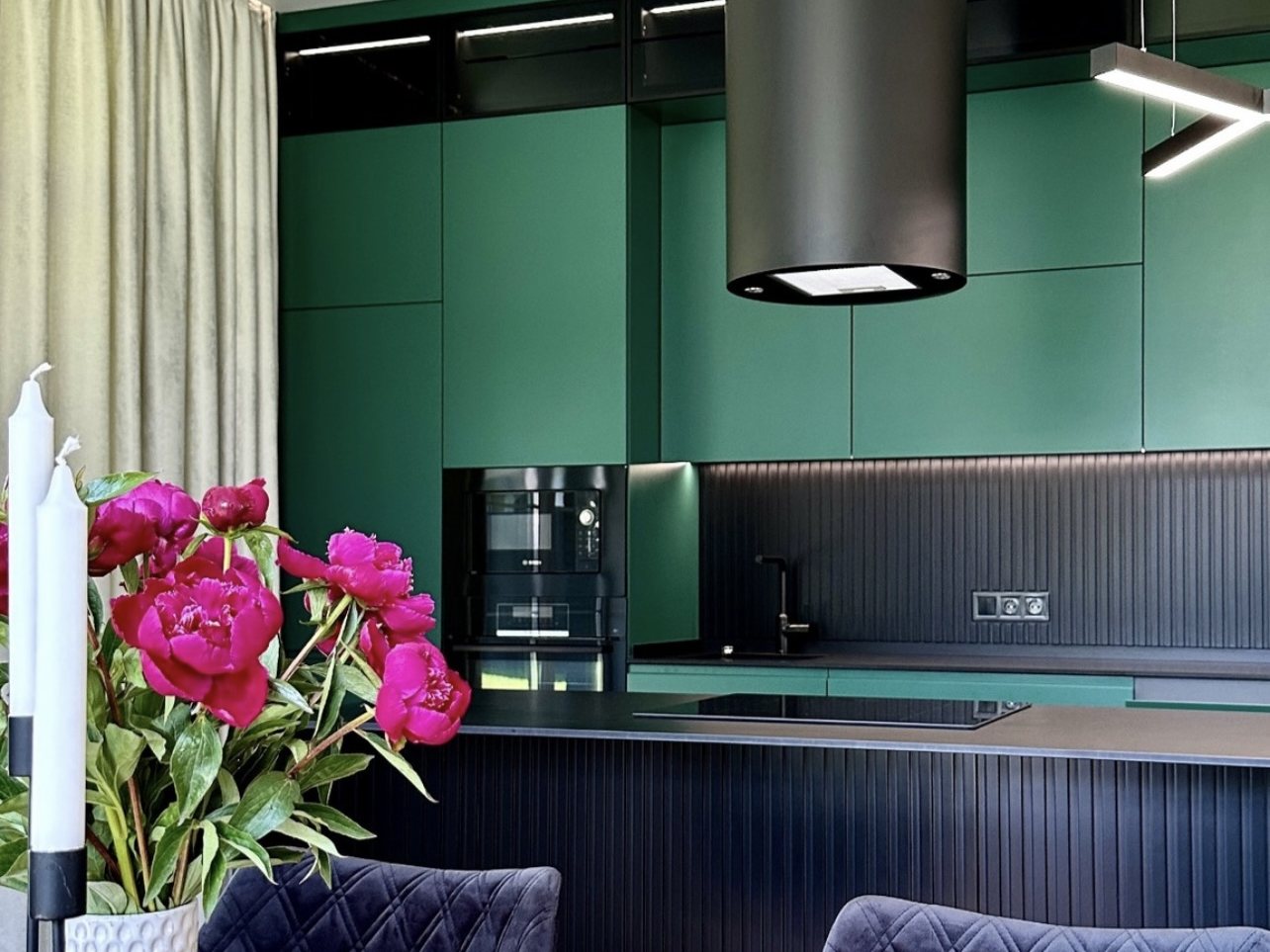 The green kitchen from the dining side: peonies on the table, a black island with a fluted front, a built-in coffee machine in the tall cabinetry and a round black hood.