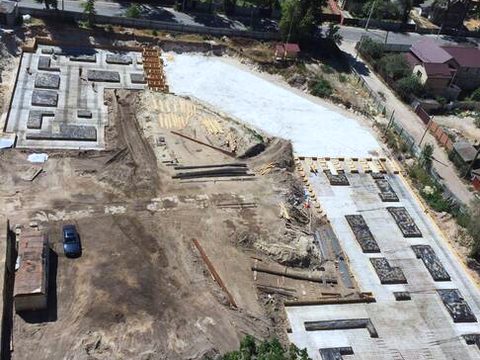 Aerial view of the site at foundation stage: the concrete foundation laid out in sections, with excavated ground and trees around it.