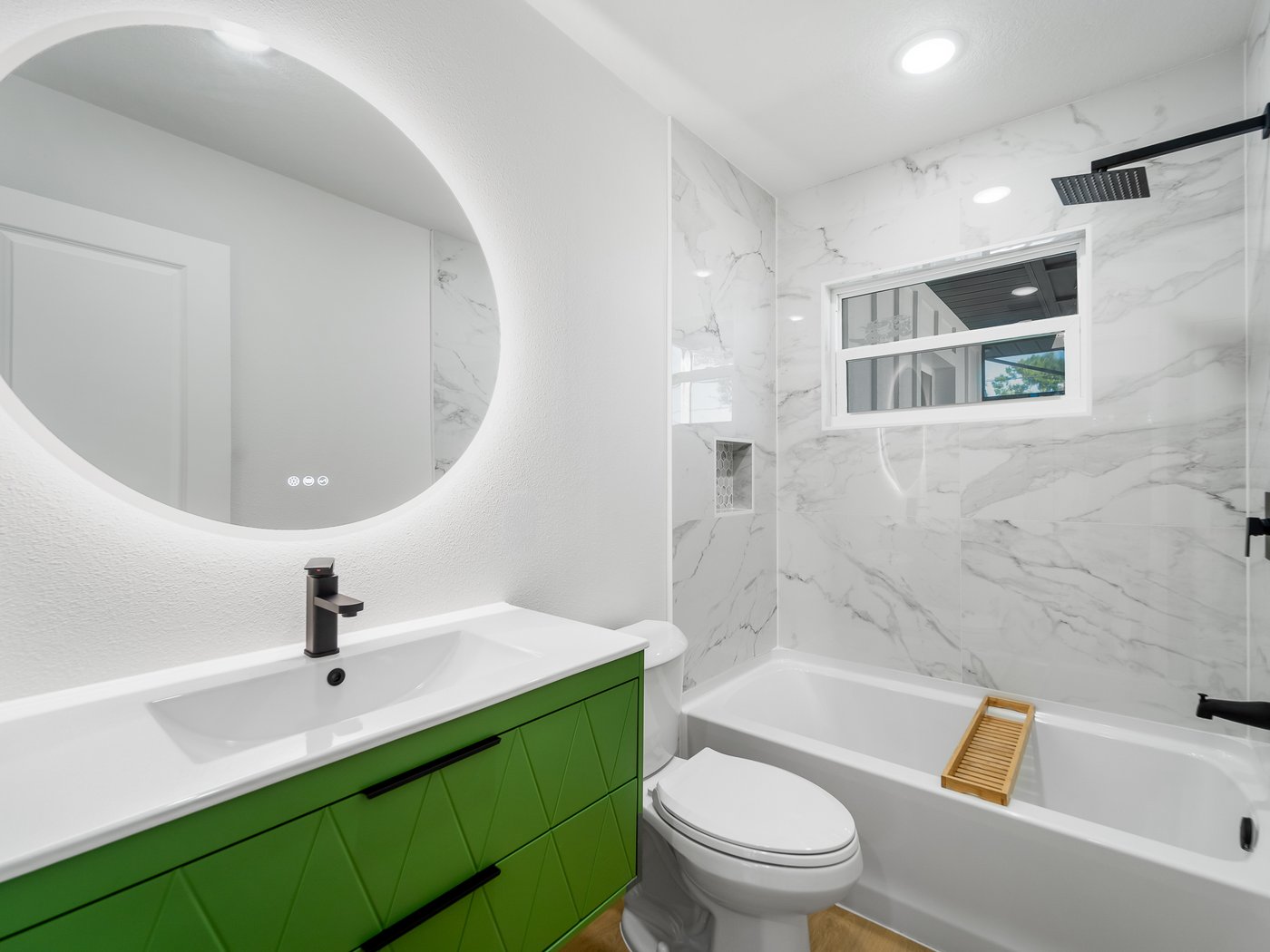 Finished bathroom with a bold green vanity, oversized round backlit mirror, and marble-tiled tub and shower.