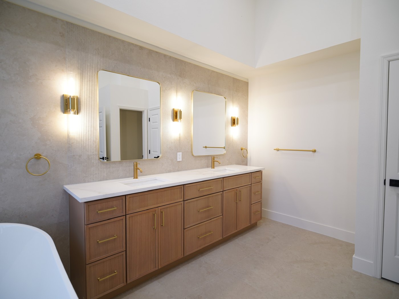 The primary bathroom vanity wall: a long stone top with twin basins, two framed mirrors between brass sconces, and the freestanding tub at the edge of the frame.
