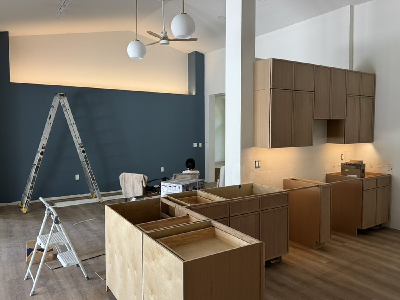 The kitchen mid-rebuild: cabinet boxes set and levelled along both walls, the island framed with no countertop yet, a ladder against the navy feature wall.