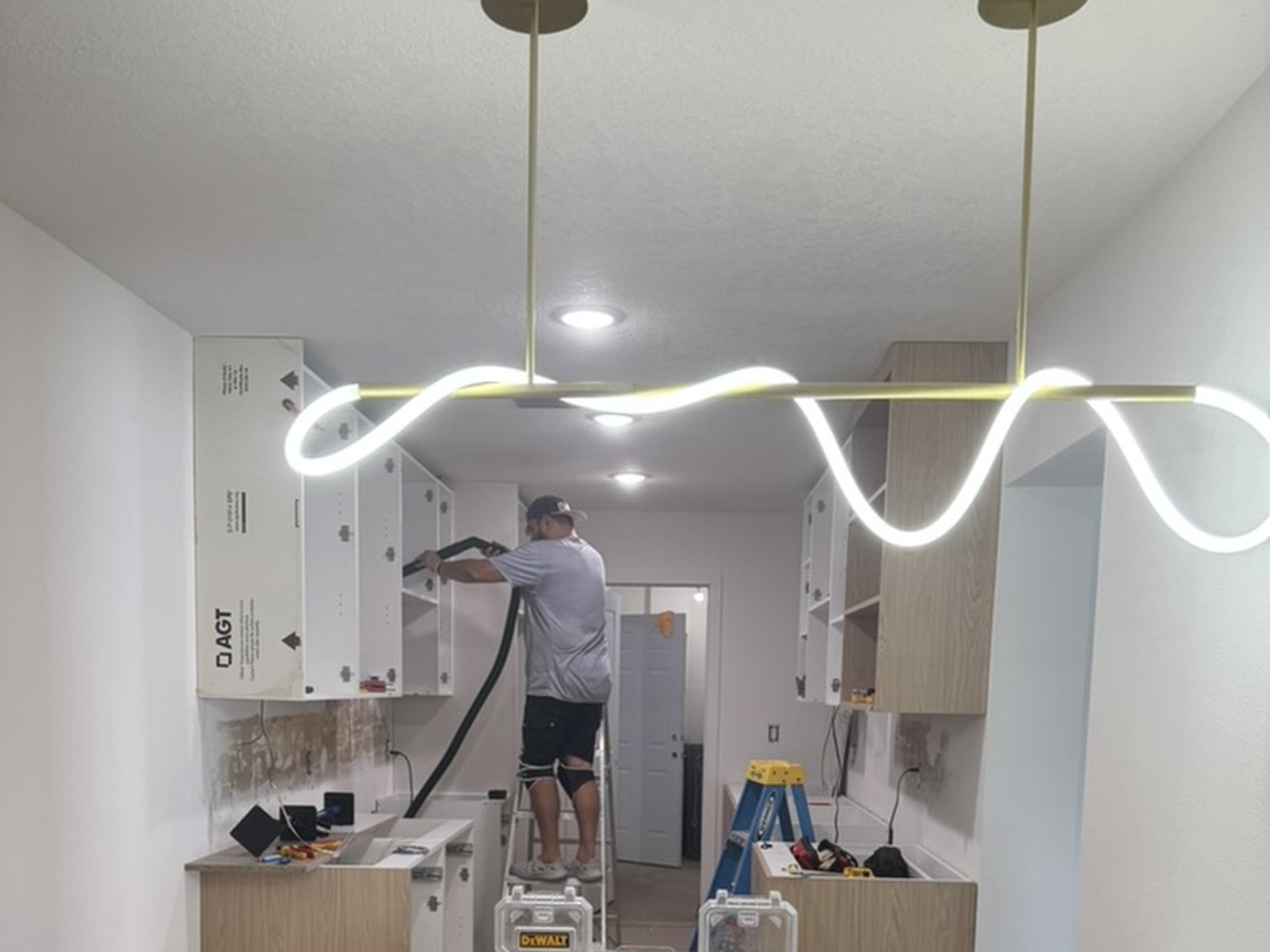 Worker installing a sculptural wavy light fixture above a kitchen island mid-construction, cabinetry not yet finished.