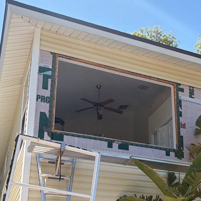 A second-floor window opening with the old unit out — siding stripped back to the housewrap, bare timber framing showing at the head and jambs, the room and its ceiling fan visible through the hole.