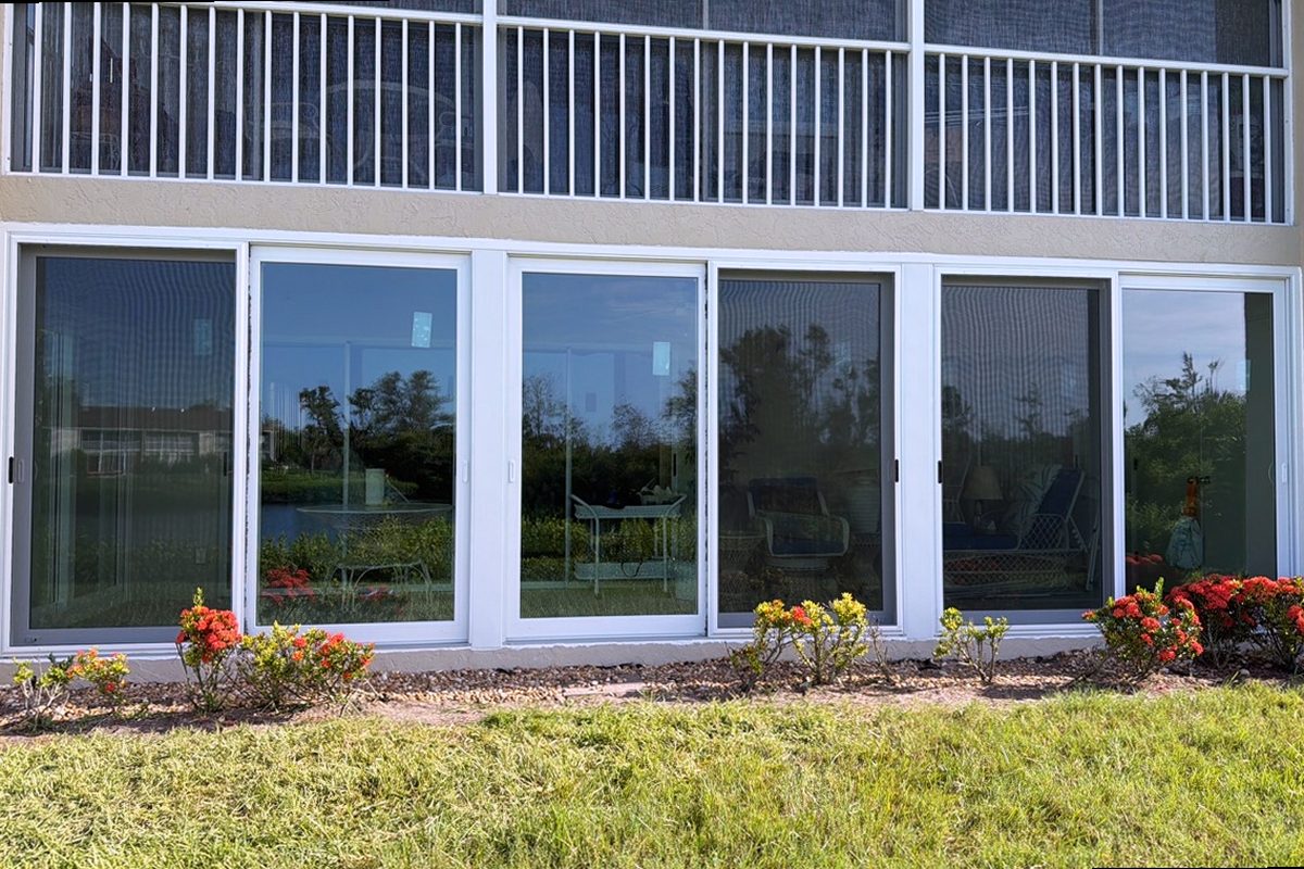The ground floor of a house replaced end to end: a run of impact windows and sliding doors under a balcony, planting beds and red flowers along the wall.