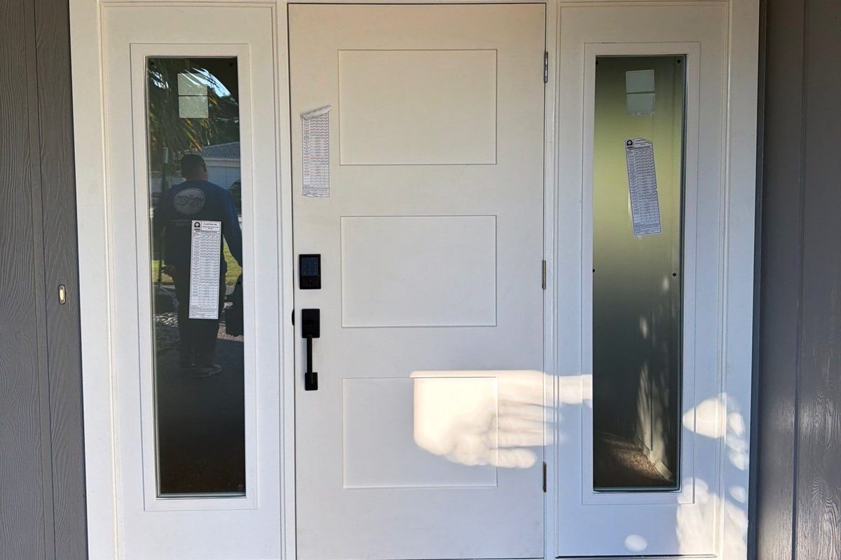 A white impact entry door with a glazed sidelight either side, set in a covered entry, the manufacturer’s paperwork still taped to the glass.
